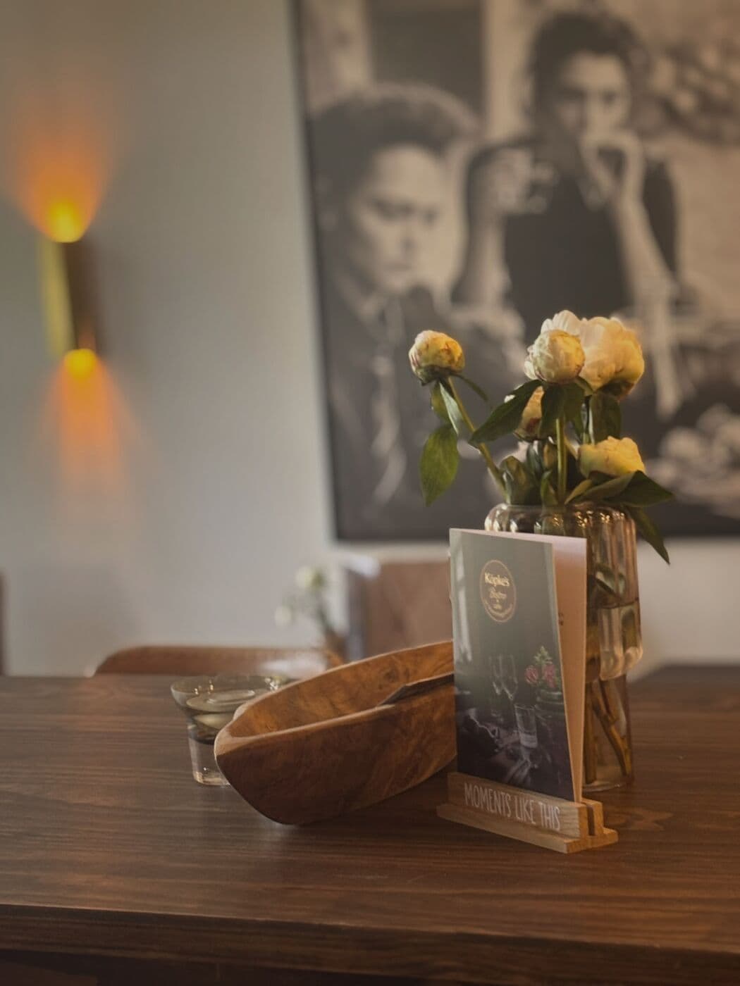 Helle Blumen in der Vase, Holzschale und Tischkarte auf einem Holztisch, dahinter eine Messing-Wandleuchte